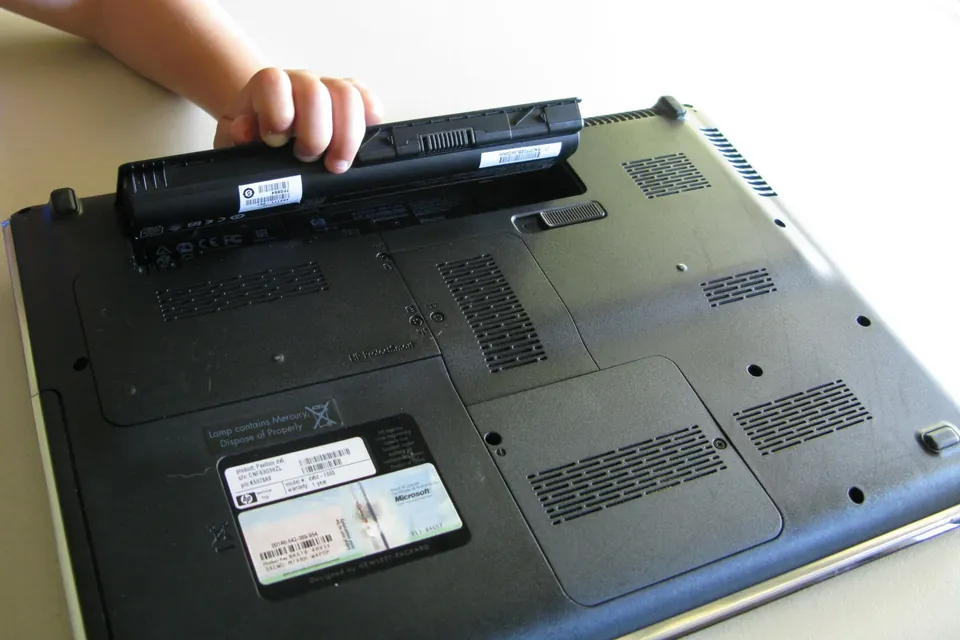 A hand lifts a removable battery from the underside of an older HP Pavilion laptop.